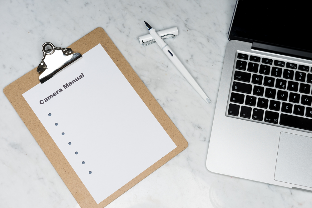 Image of a marble desktop with a pen, laptop, and clipbard. The clipboard has a sheet of paper with the words Camera Manual at the top followed by a list of bullets arranged vertically.